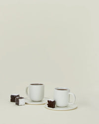 The Simple Porcelain Espresso Cups & Saucers - Set of 2 by Hawkins New York are shown on a plain light background, each on its saucer and paired with a chocolate brownie and a sugar-dusted brownie.