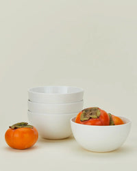 Four Simple Porcelain Large Bowls by Hawkins New York are stacked next to a white bowl filled with three persimmons, with another persimmon in front, all set against a plain, light background.