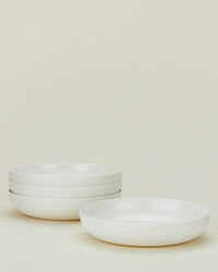 Four plain white shallow bowls from the Hawkins New York Simple Porcelain Dinnerware - 16 Piece Set, three stacked on the left and one placed separately on the right, set against a light beige background.