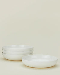 Four Simple Porcelain Low Bowls - Set of 4 from Hawkins New York are stacked on the left, with one more bowl in front right, all set against a light cream background—ideal for displaying elegant porcelain dinnerware.