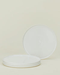 Four round, white plates from the Hawkins New York Simple Porcelain Dinnerware 16 Piece Set are arranged on a neutral background—three stacked and one upright to show its smooth, plain surface.