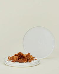 A small pile of thin, crispy chips sits on a fine plate beside two empty Hawkins New York Simple Porcelain Salad Plates from the set of 4, all arranged against a plain, light background.