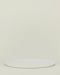 The Simple Porcelain Serving Platter by Hawkins New York, a round white porcelain piece, rests on a light beige background with empty space above, reflecting the brand’s minimalist elegance.