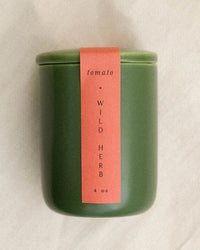 A green ceramic jar from LEIF with a red Tomato & Wild Herb label (4 oz), placed vertically on the front, is shown on a light background—perfect for mini candles made from a calming soy wax blend.