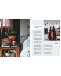 A Wild Creations candle from Simon & Schuster burns on a wooden nightstand next to books, a basket, indoor plants, and a magazine page featuring a woman smiling in front of a CANDLE CO. sign.