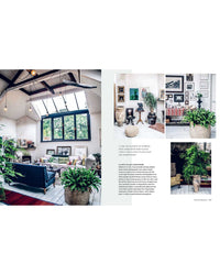 A bright, spacious living room with large windows and modern furniture is decorated using ideas from Wild Interiors by Simon & Schuster—lush house plants enhance the space and complement a gallery wall of framed art and photos.