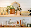 A wooden shelf displays potted plants, patterned pillows, glassware, candles, books, and a small mirror against a white shiplap wall.