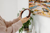 A person wearing a beige robe holds a round mirror in front of a plant and a colorful painting on a wall.