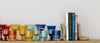 A row of colorful glassware and several upright books held by stone bookends are arranged on a wooden shelf against a plain white wall.