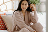 A woman with long dark hair sits on a bed, wearing a matching light-colored knit sweater and pants, with cushions and a plant in the background.