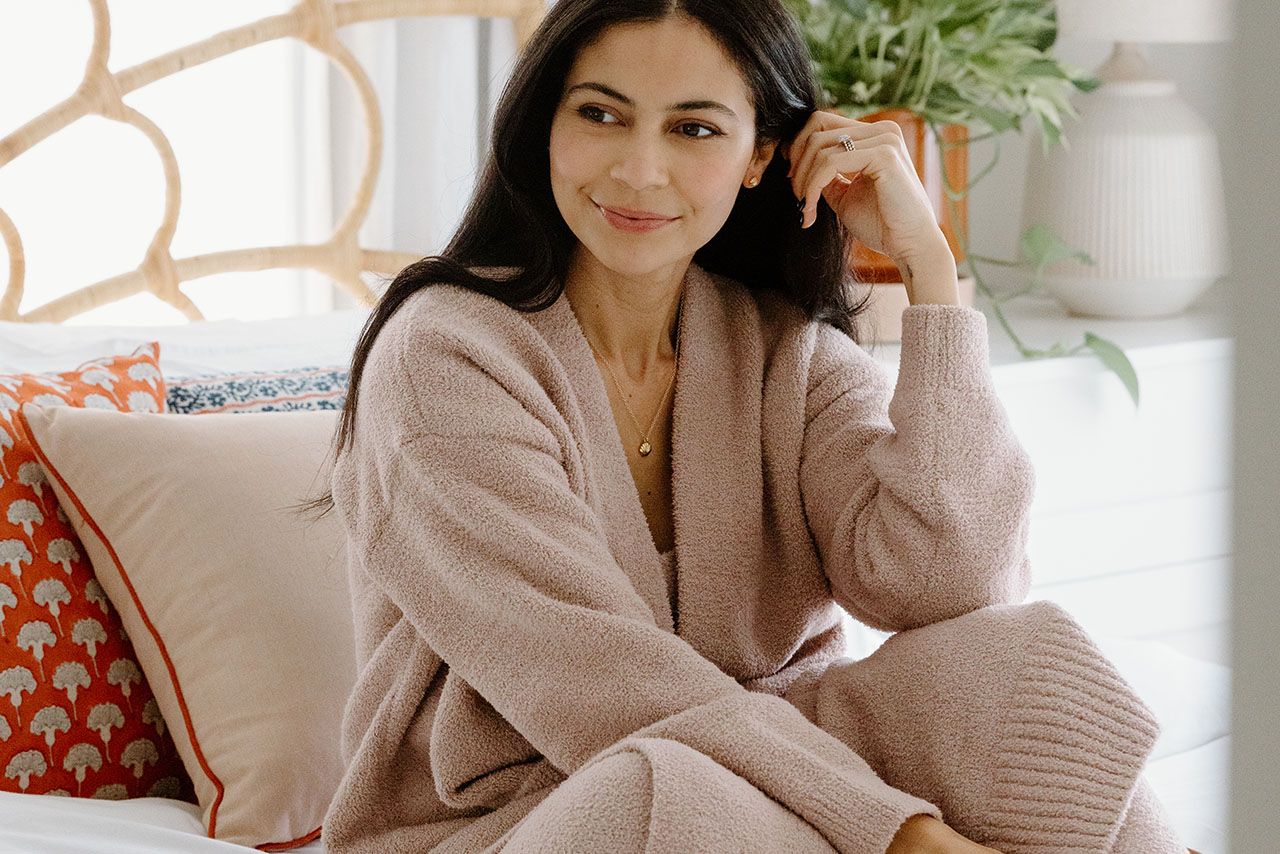 A woman with long dark hair sits on a bed, wearing a matching light-colored knit sweater and pants, with cushions and a plant in the background.