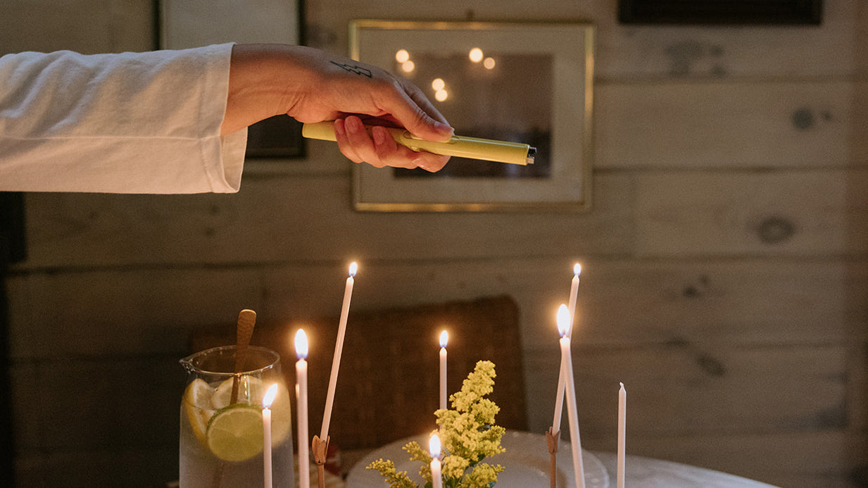 A person uses a lighter to light candles on a table set with a floral arrangement and a glass pitcher of water with lemon slices.