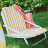 A yellow and white striped lawn chair sits on grass with a pink tote bag and a drink container resting on it, surrounded by green plants.