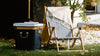 A striped lounge chair with a beige throw sits on grass next to a cooler and a book, with a small tree and white stones in the background.