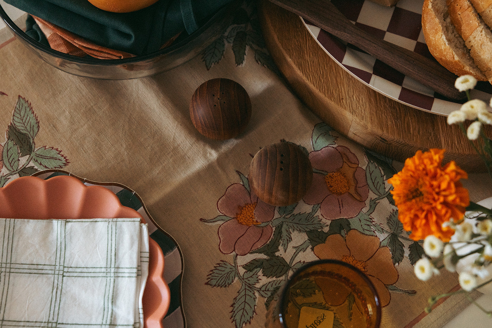 A tabletop with floral-patterned cloth, two wooden salt and pepper shakers, stacked plates with a plaid napkin, a loaf of bread, orange marigolds, and an amber glass.