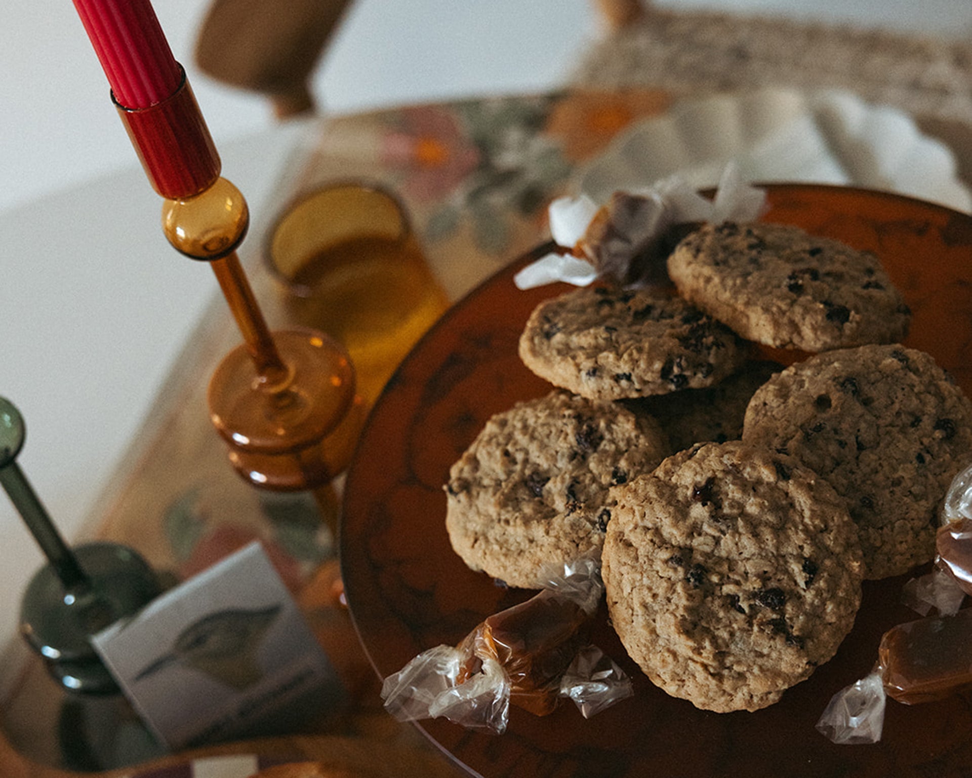 A plate of chocolate chip cookies and wrapped candies sits on a table with amber glassware, a red candle in a holder, and a patterned tablecloth.