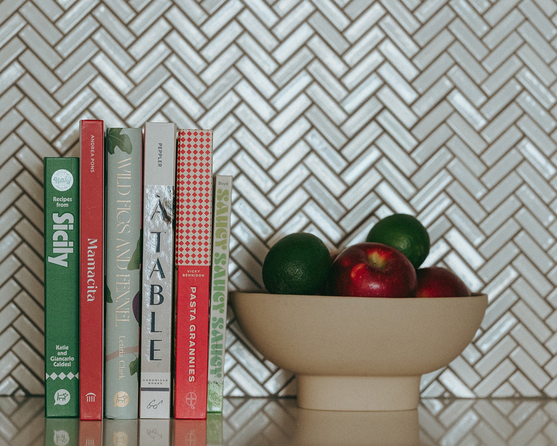 A row of colorful cookbooks stands upright next to a beige bowl filled with red apples and green limes, set on a kitchen counter with a white herringbone tile backsplash.