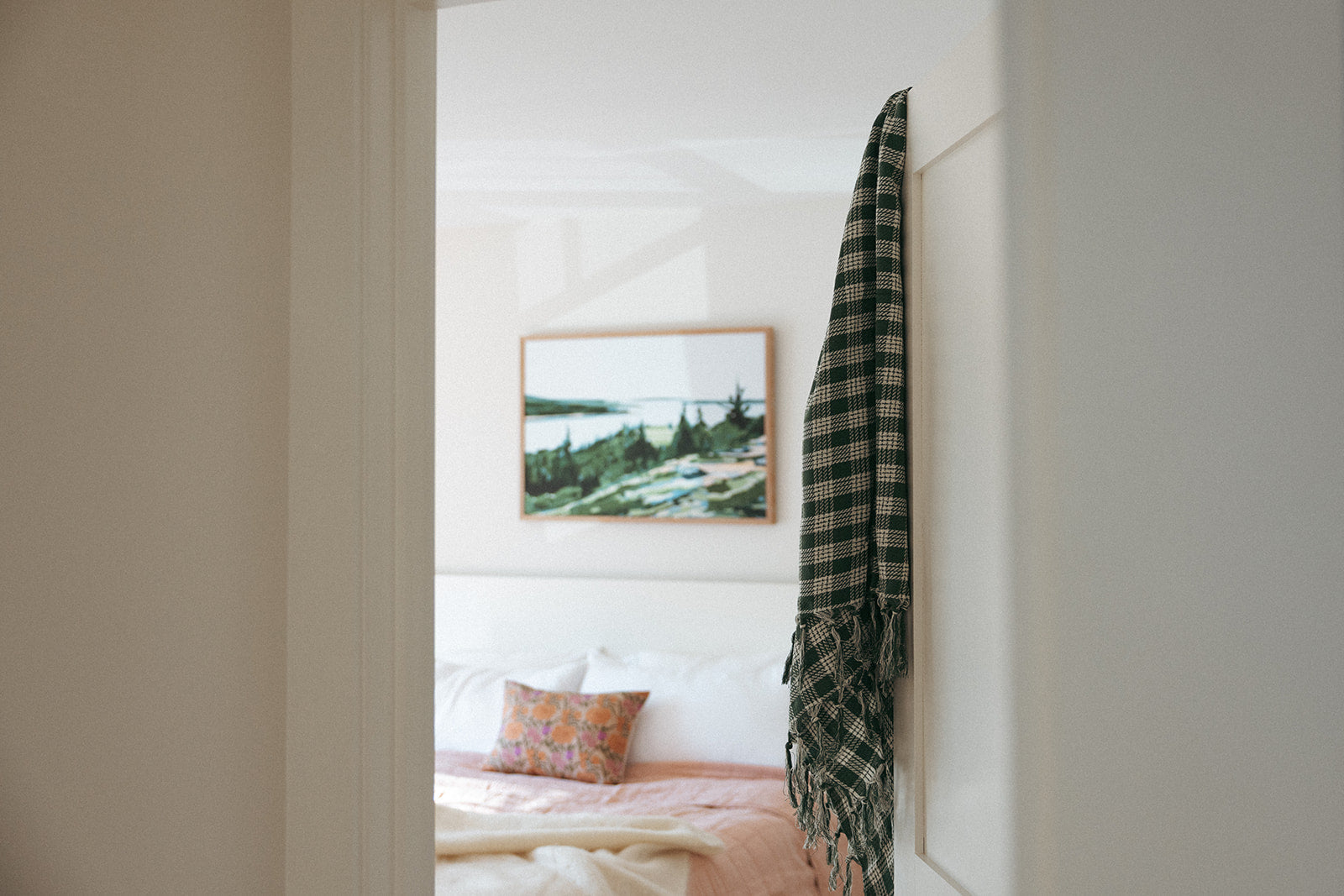 A plaid scarf hangs on a white door with a cozy bedroom in the background, featuring a bed with a pink blanket, a decorative pillow, and a landscape painting on the wall.