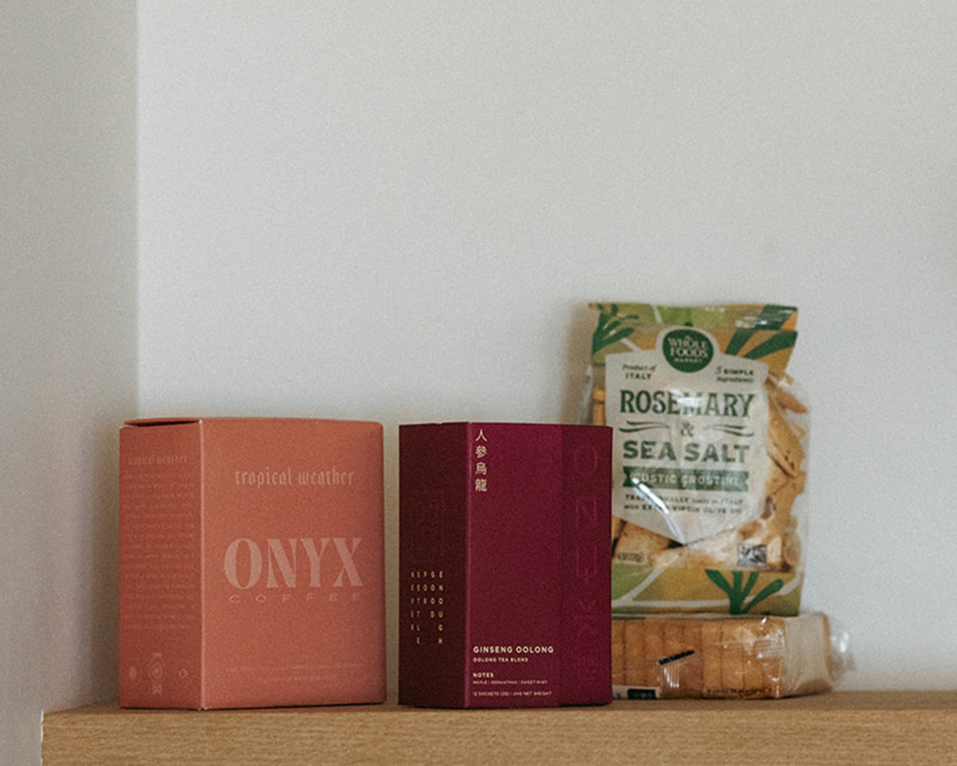 Two boxes and a bag of snacks are arranged on a wooden shelf against a white wall. The items include Onyx Coffee, Ginseng Oolong tea, and rosemary sea salt crackers.