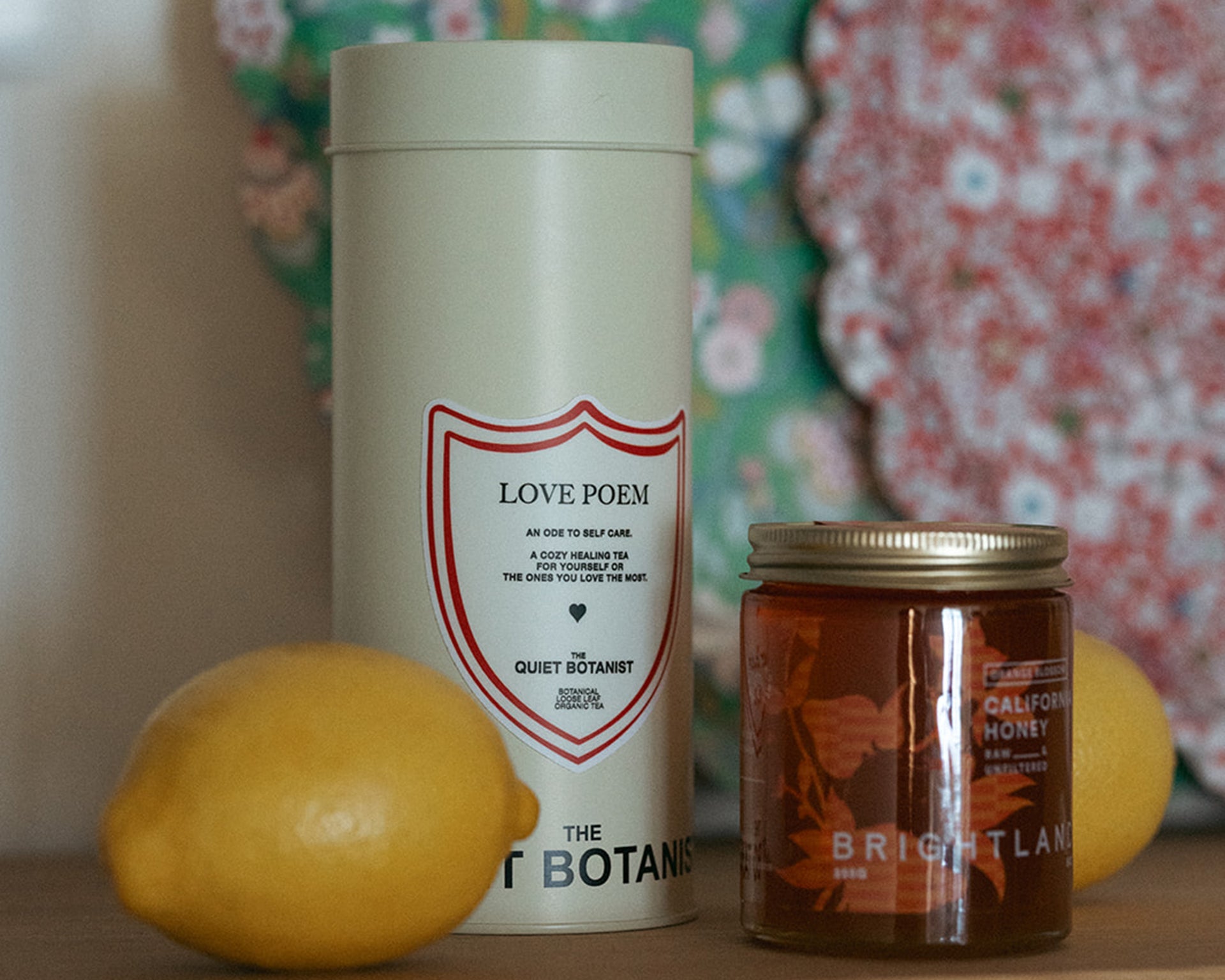 A tall tin labeled Love Poem stands next to a jar of Brightland honey and two lemons, with a floral-patterned object blurred in the background.