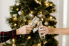 Two people clink champagne glasses in front of a decorated Christmas tree with lights and ornaments. One wears a plaid shirt, the other has red nail polish.