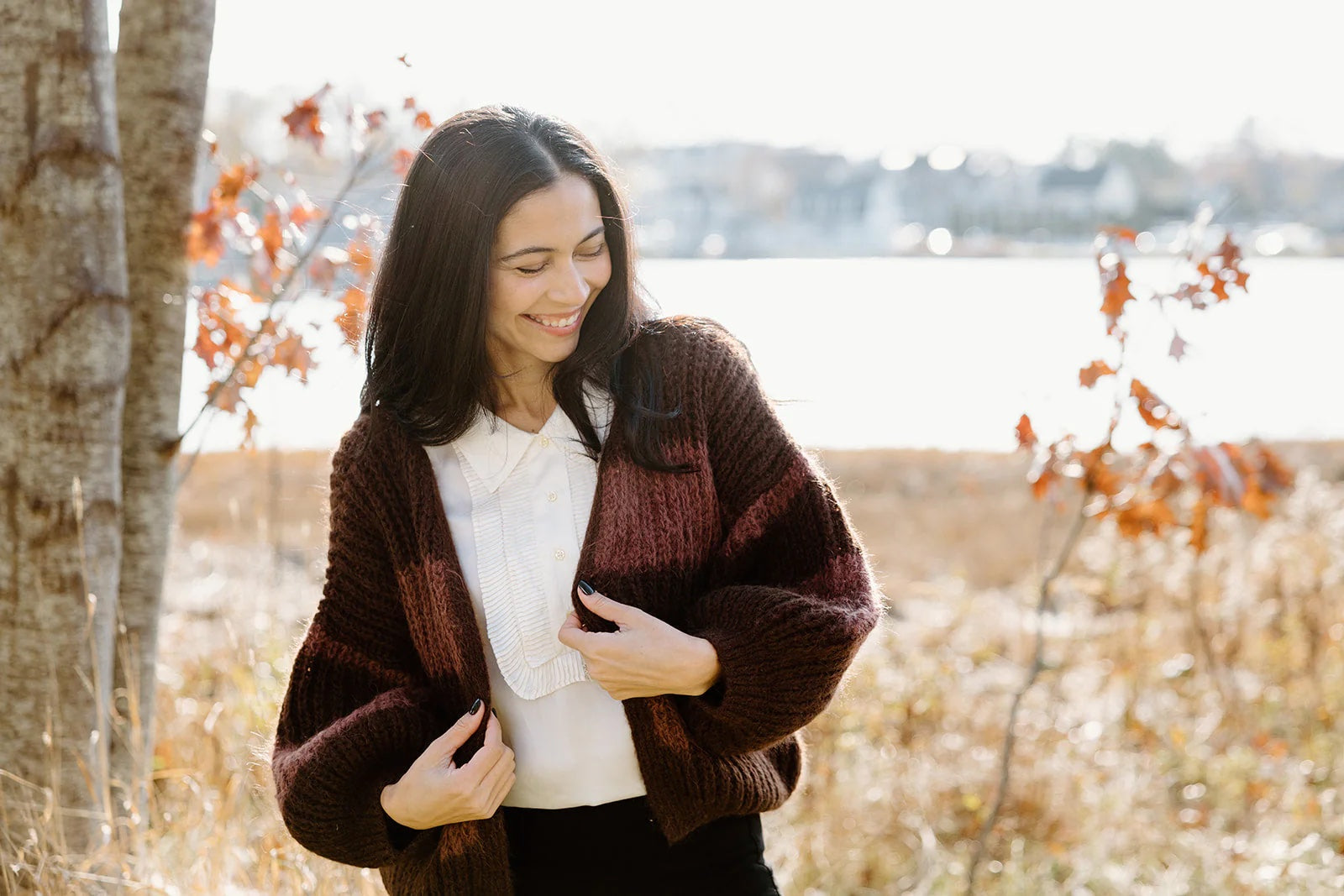A woman wearing a white blouse and a brown knit cardigan stands outdoors near trees and autumn leaves, smiling and looking down.