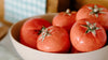 A beige bowl filled with five shiny, red tomatoes with green stems, placed indoors near a checkered cloth in the background.