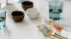 A neatly set table with green glasses, small bowls, a white plate, gold cutlery, and napkins with a decorative orange accent.