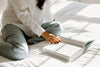 A person in a white shirt and jeans sits cross-legged on a bed, reading an open book in natural light.