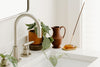 A kitchen sink with a metal faucet, a potted plant, a brown ceramic pitcher, and a burning incense stick in a holder on the white countertop.