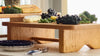 A wooden serving board on a table holds grapes, cheese, green beans, and leafy greens. Plates and utensils are arranged nearby.
