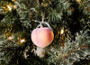 A peach-shaped ornament hangs on a green Christmas tree with string lights in the background.