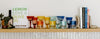 A wooden shelf displays colorful glassware, books, a bottle, and a small plant against a white wall with a herringbone tile backsplash below.