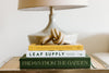A stack of three books topped with a decorative knot sits on a wooden table beside a beige lamp.