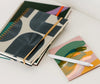Three notebooks with abstract covers, stacked on a white surface next to a small notepad with colorful brush strokes and a white pen.
