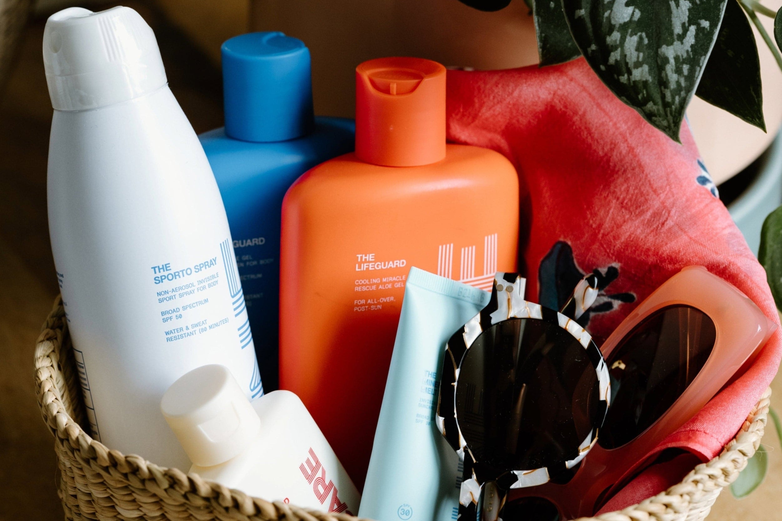 A woven basket contains sunscreen bottles, a tube, a pair of sunglasses, and a coral-colored cloth next to a leafy plant.