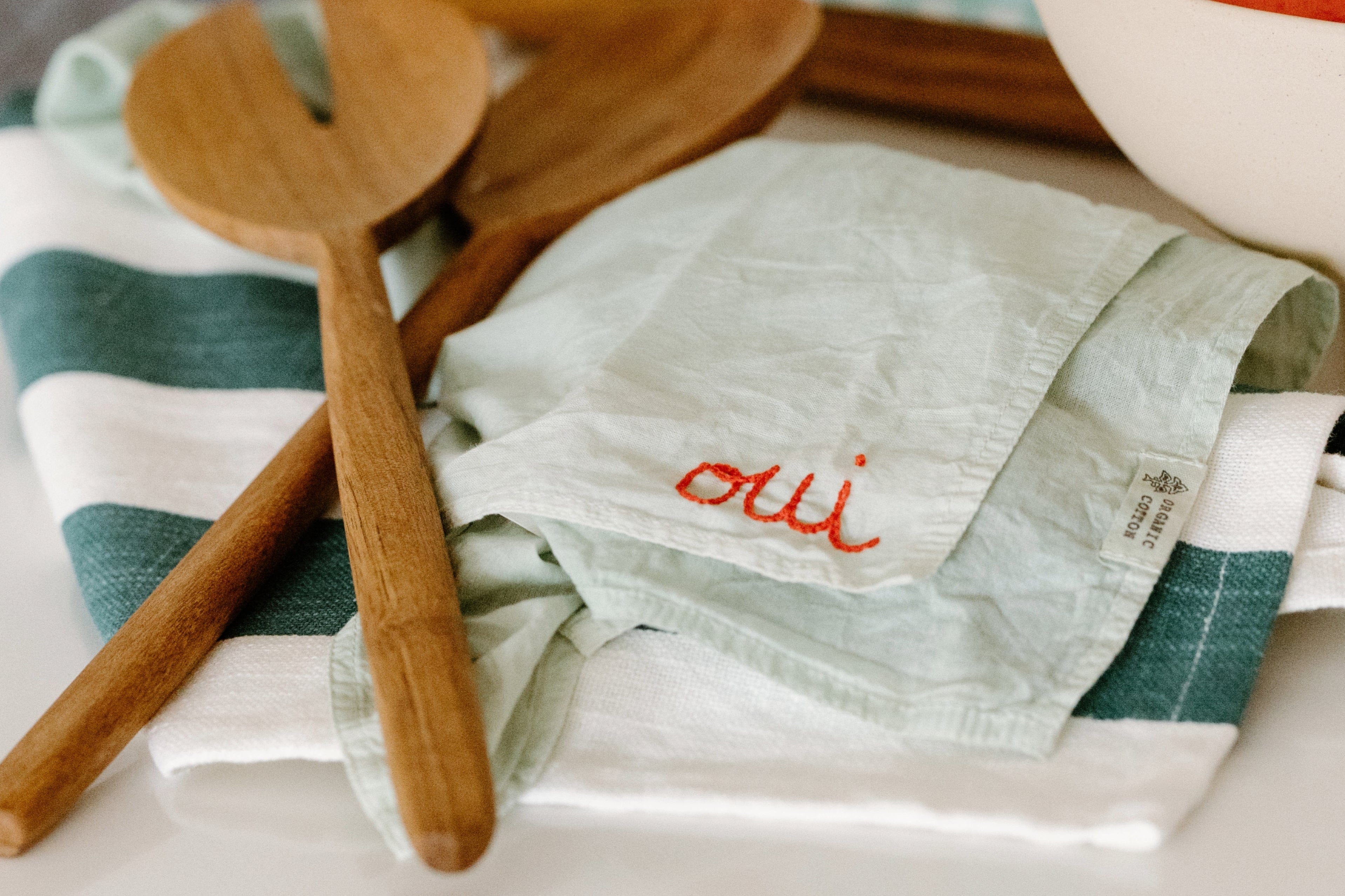 Two wooden cooking utensils rest on a green-and-white striped cloth and a light green napkin embroidered with the word oui in orange.