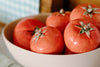 A beige bowl filled with five ripe red tomatoes, each with green stems, placed on a light surface with a checkered cloth in the background.