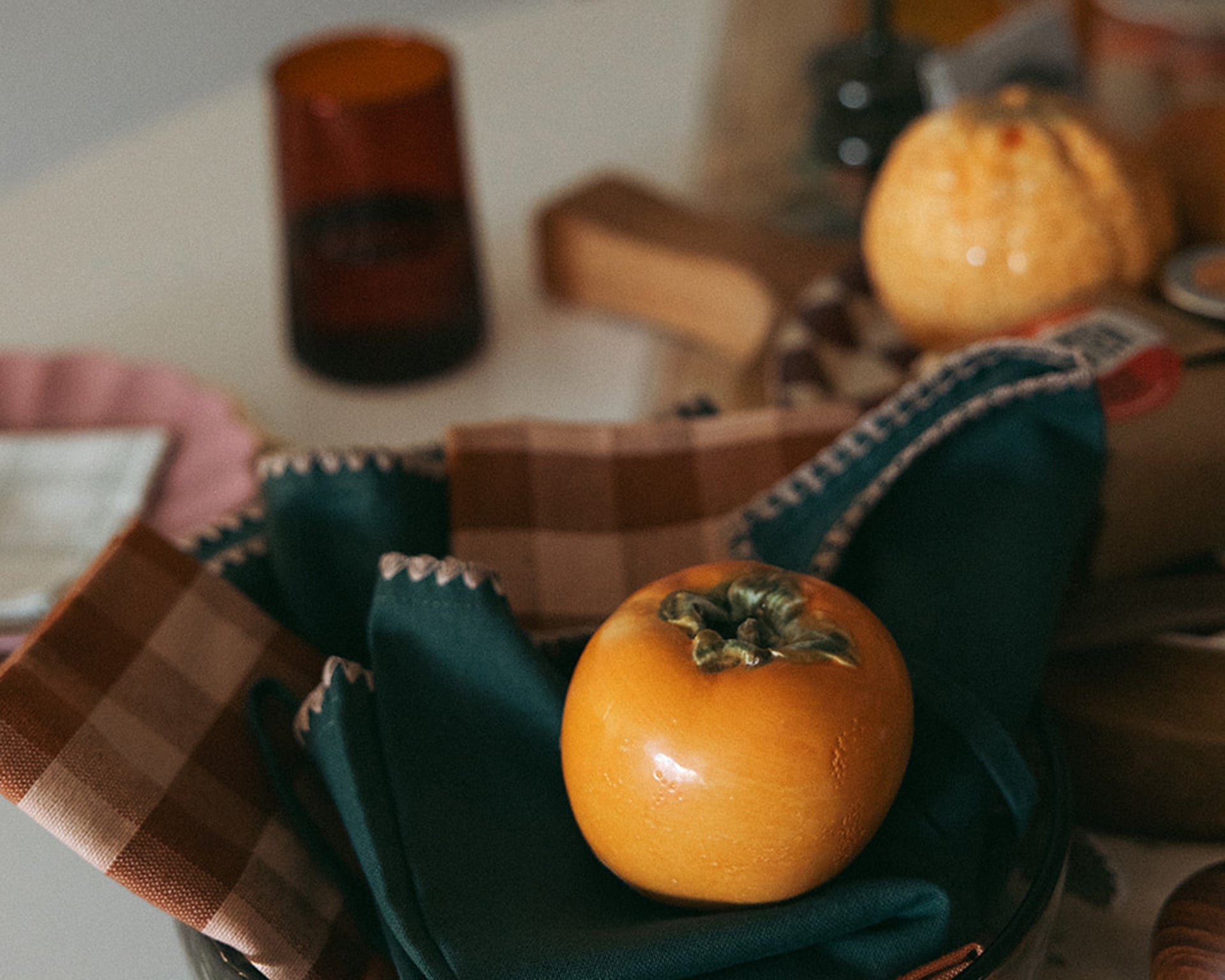 A beige bowl filled with five ripe red tomatoes, each with green stems, placed on a light surface with a checkered cloth in the background.