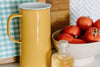A yellow metal pitcher, a clear bottle of liquid soap, and a bowl of tomatoes on a kitchen counter with a patterned towel in the background.