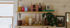Two wooden shelves display books, canned goods, bottles, glassware, candles, a potted plant, and a framed landscape photo against a light wooden wall.