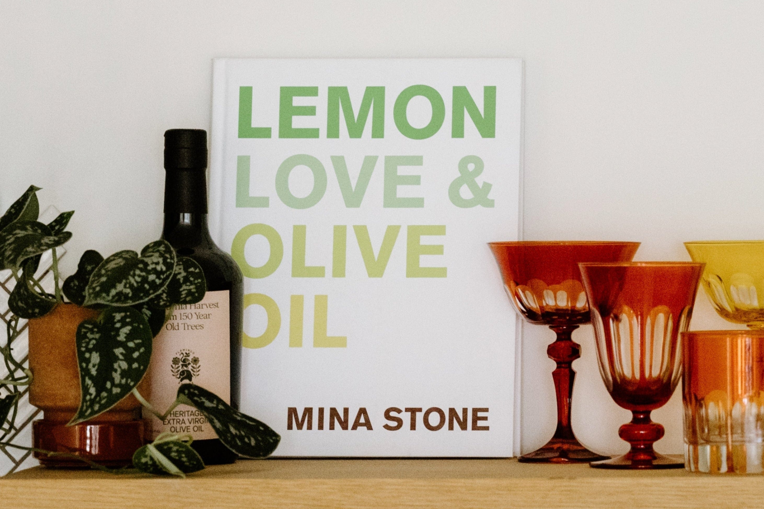 A cookbook titled Lemon Love & Olive Oil by Mina Stone is displayed upright next to a bottle of olive oil, amber glassware, and a small potted plant.
