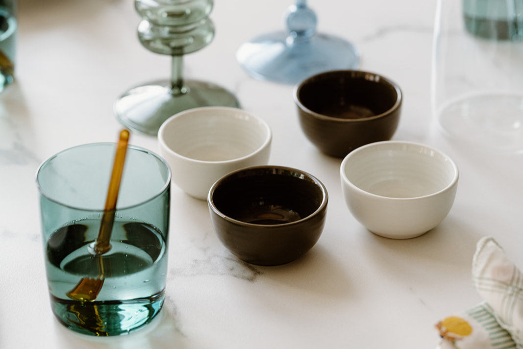 Four small ceramic bowls, two black and two white, arranged on a white table next to a green glass cup with an amber stirring stick and glassware in the background.