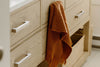 An orange woven towel hangs over the handle of a light wood bathroom cabinet with a wicker basket below.