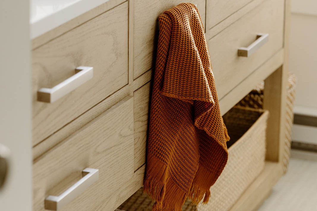 An orange woven towel hangs over the handle of a light wood bathroom cabinet with a wicker basket below.