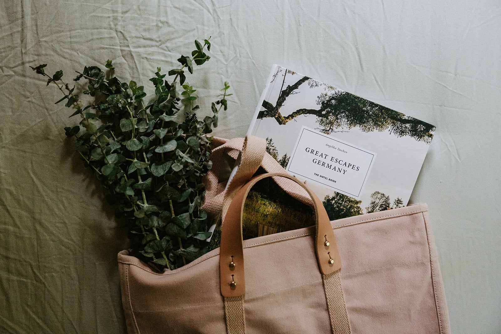 A beige tote bag containing green foliage and a book titled Great Escapes Germany rests on a light-colored fabric surface.