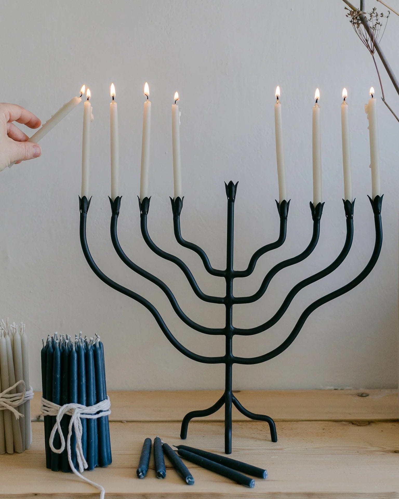 A hand lights the final candle on a black metal menorah with all candles burning. Bundles of blue and white candles rest on a wooden table beside the menorah.