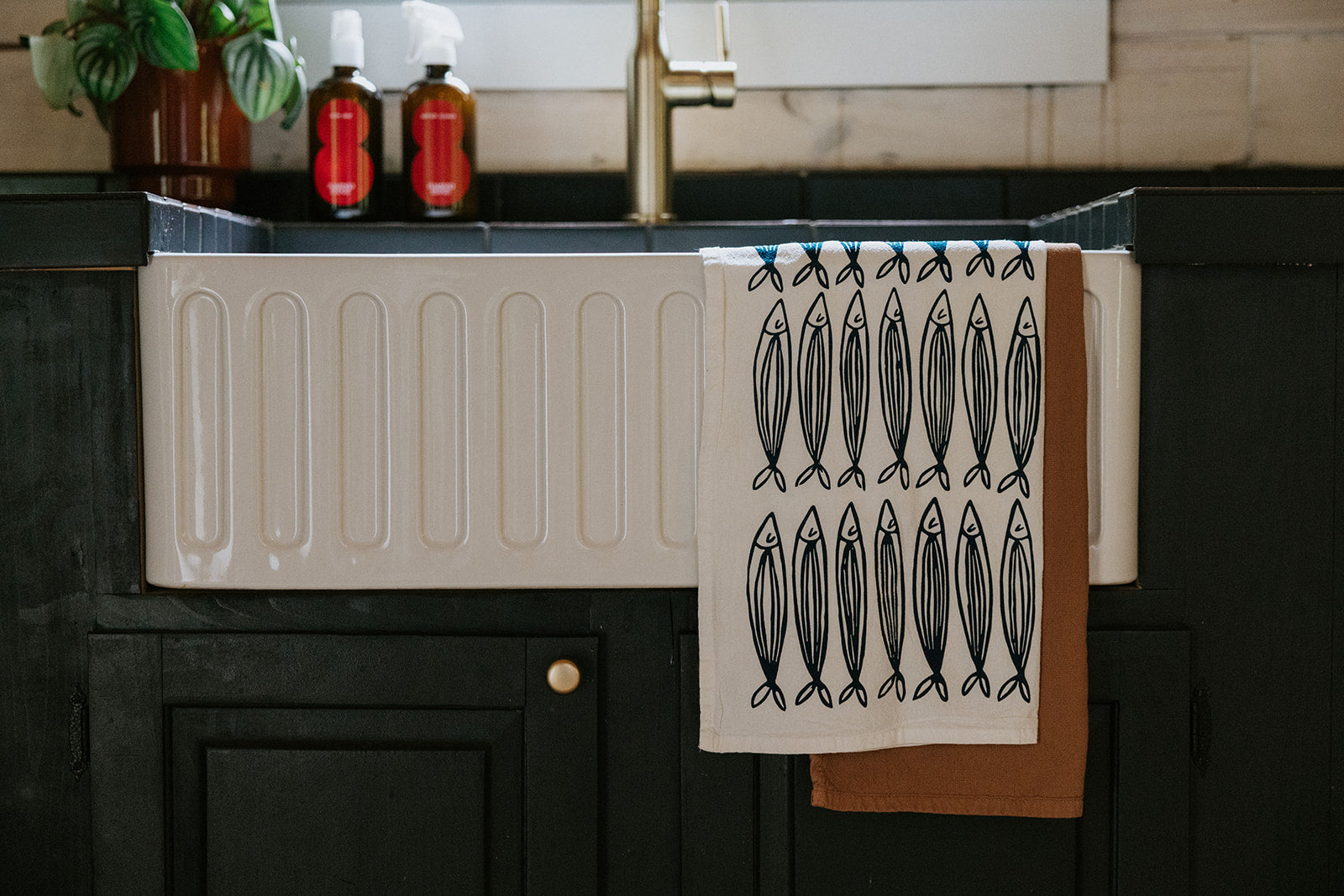 A kitchen sink with a tea towel featuring a fish pattern draped over the front, brown towel underneath; two bottles and a potted plant are in the background.