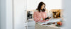 A woman stands in a modern kitchen, reading the label on a bottle while a cookbook lies open on the counter in front of her.
