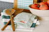 Two wooden utensils rest on striped dish towels beside a beige bowl filled with tomatoes. A light green cloth with oui embroidered in red is on top of the towels.